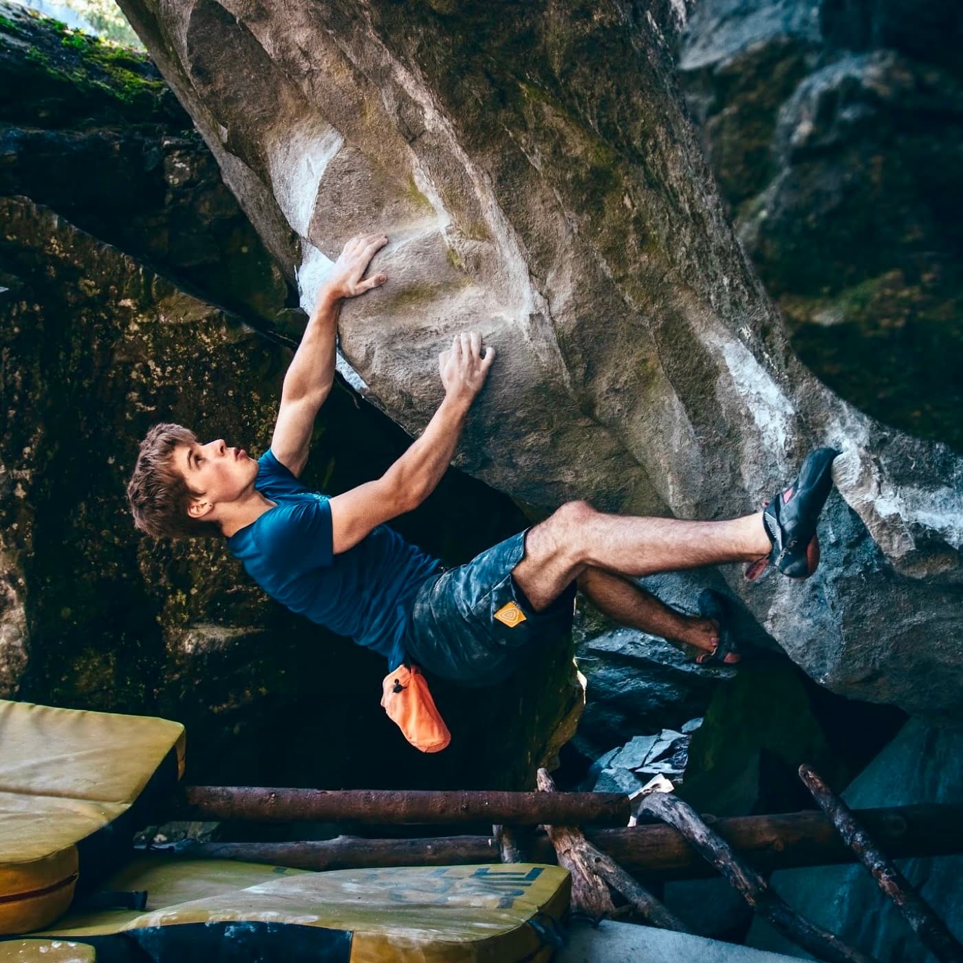 Bouldering out from under a steep grey roof in the forest, crash pads on the ground below